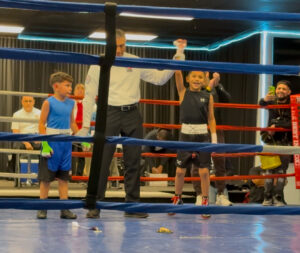 A Tucson youth boxing gym scene featuring kids training in self-defense techniques. The image includes a young boxer practicing with a coach, working on footwork and blocking skills in a professional gym setting.